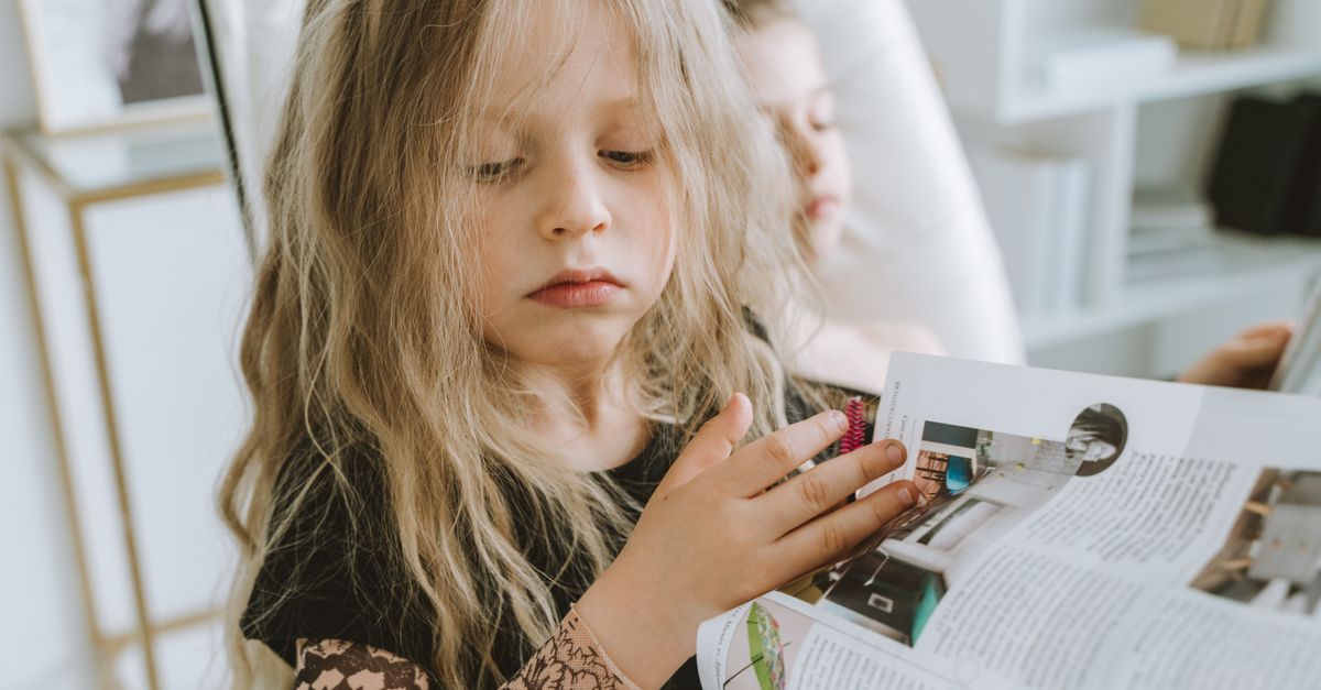A young girl with long blonde hair attentively reading a magazine indoors.