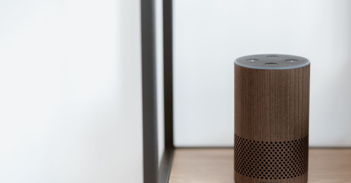 A sleek, modern smart speaker displayed on a wooden shelf in a minimalist room setting.