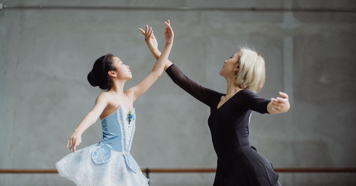 Low angle side view of graceful Asian student in tutu dancing with teacher with raised arms in ballet class against wall